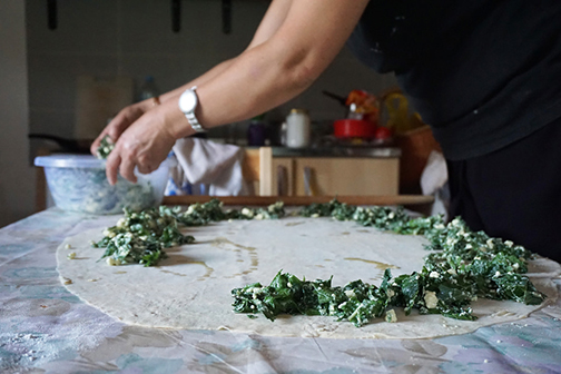Spreading a mixture of greens and cheese on top of rolled-out burek dough for pita in Bosnia.